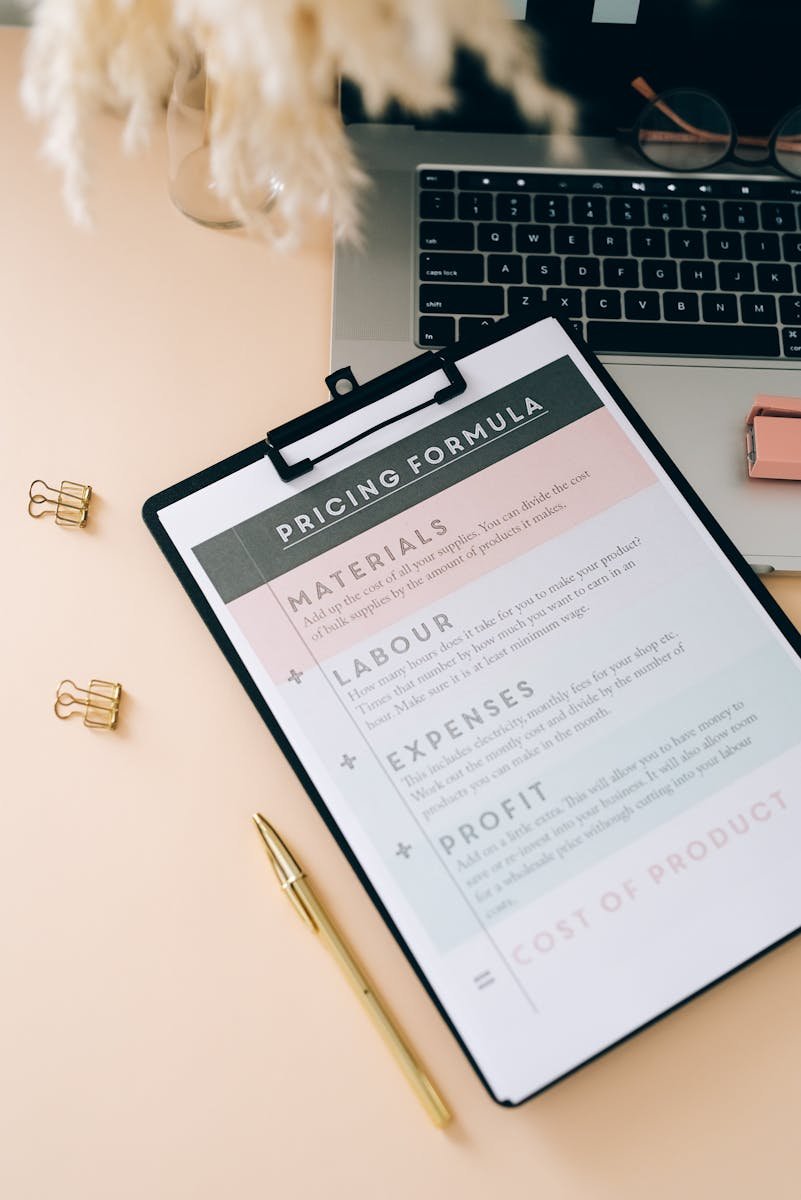 Organized workspace featuring a pricing formula document, laptop, pen, and clips on a desk.