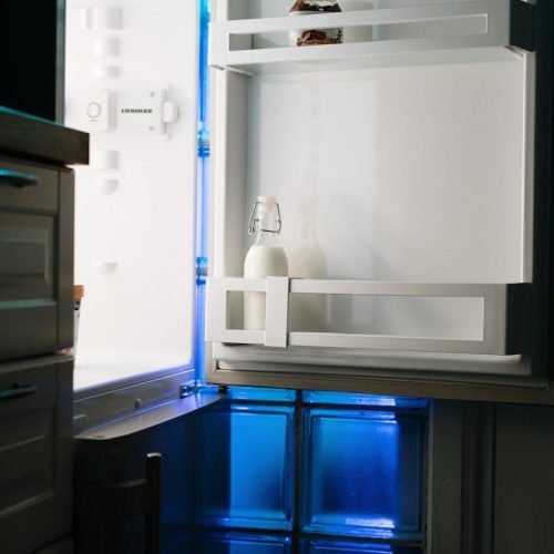 An open refrigerator with minimal contents illuminated by neon light in a home kitchen.